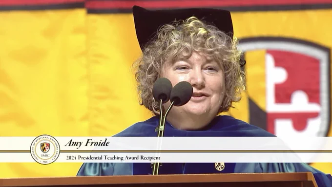 A female professor with short, curly hair stands at a podium in higher education regalia, with lower-third reading- "Amy froide. 2024 presidential teaching award recipient" 