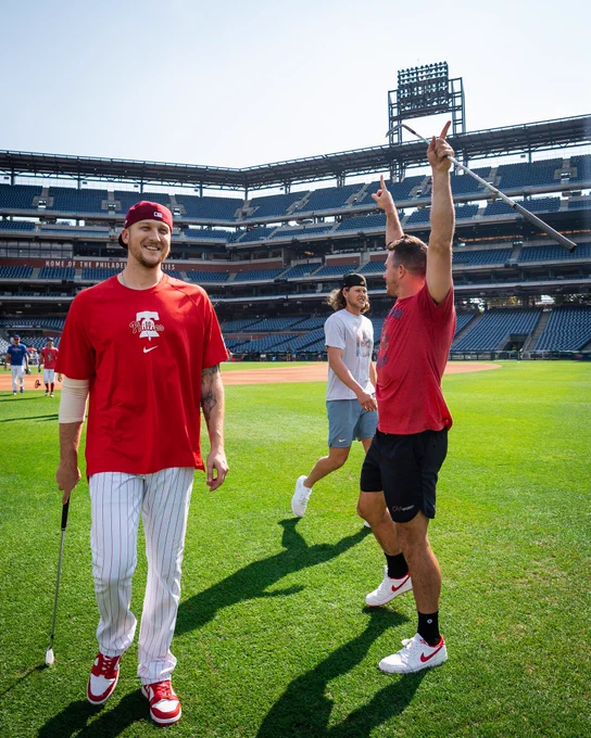 J.T. Realmuto celebrates winning the closest to the pin competition. He is wearing a red shirt and black shorts. Jeff Hoffman stands next to him laughing. Both are holding gold clubs.