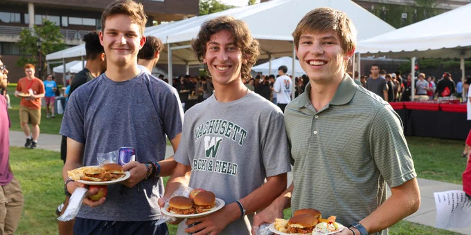 Three students holding plates of food at RPI Welcome Fest, smiling at the camera, with a tent and other attendees in the background.