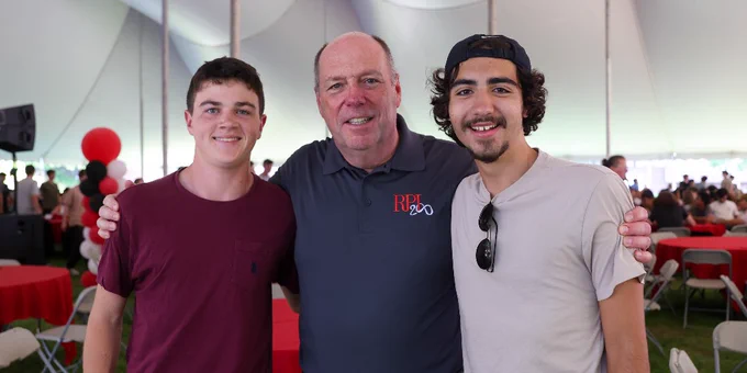 RPI President Martin Schmidt poses with two students under a large tent. They are smiling and there are red balloons and tables in the background.