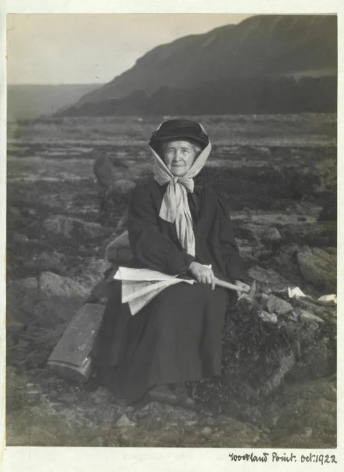 Archival photo of Elizabeth Anderson Gray sitting in a moorland area. She's holding a hammer-like pickaxe against some rocks. She's wearing a long dark overcoat and a dark hat, wrapped with a light scarf tied in a bow beneath her chin.