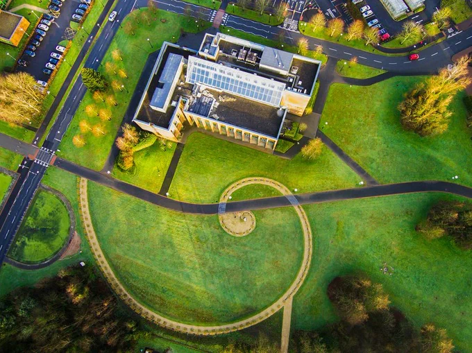 An aerial photo looking down on a vast lawn with pathways crossing it. In the middle is a Fibonacci circle made of trees, spiraling more tightly as it reaches the centre. There is also a large white building seen from above and surrounded by trees
