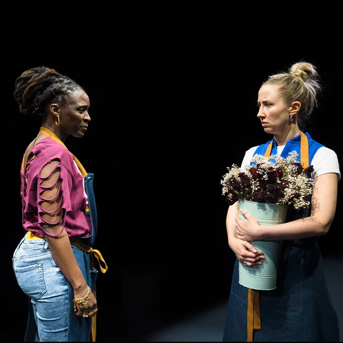 Two individuals on a dark stage, one holding a bouquet of flowers and facing the other, who is wearing a colourful  top and denim apron.
