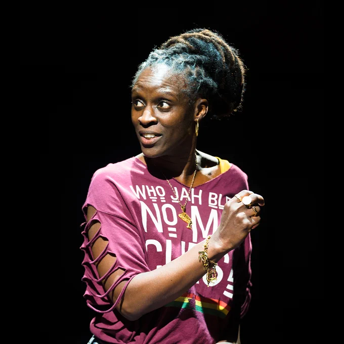 Person in a purple T-shirt speaking on stage, with focused stage lighting highlighting their expression.
