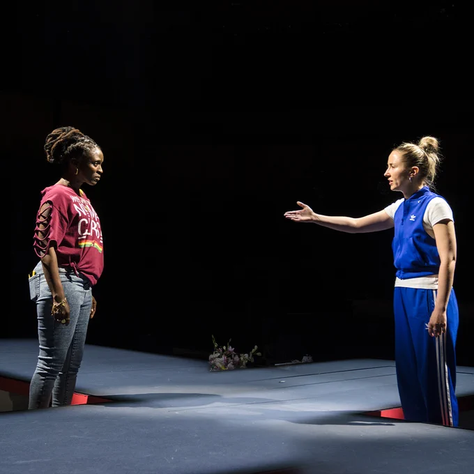 Two actors on stage in a theatrical production, one wearing purple T-shirt and the other in a blue tracksuit, interacting under a spotlight.