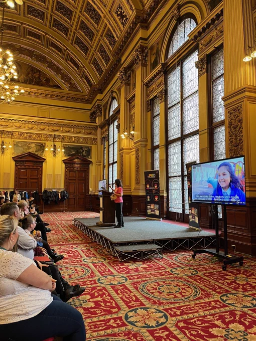A member of the living streets team is on a stage in front of an audience in an affluent looking hall at Glasgow City Chambers.  There's a screen behind the stage showing a video about WOW - the walk to school challenge. 