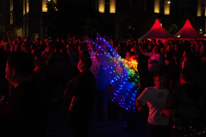 Crowd under red lighting. A person lifts their wings that light up in rainbow colours. Photo by TomPhoto.