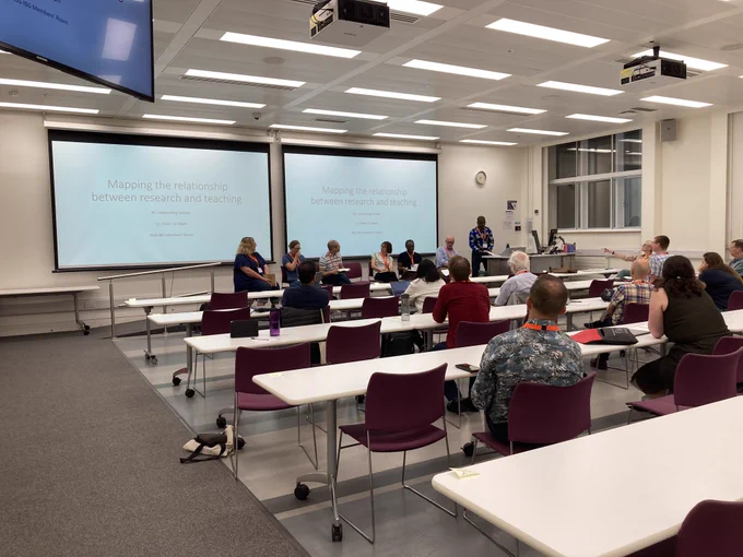 A classroom with rows of tables and chairs.  At the from facing the audience are the panel chair and six discussants.  The audience are watching the discussion