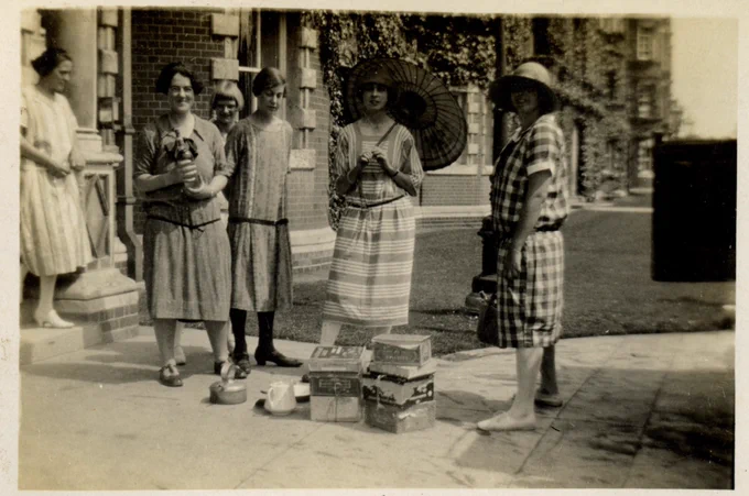 Sepia photograph of six female students standing outside the Founder's Building in 1920s summer dresses. One of them is holding a parasol and another is wearing a big sun hat. Another student is holding a bottle. On the ground in front of them are two piles of tin boxes, a teapot and a metal kettle.