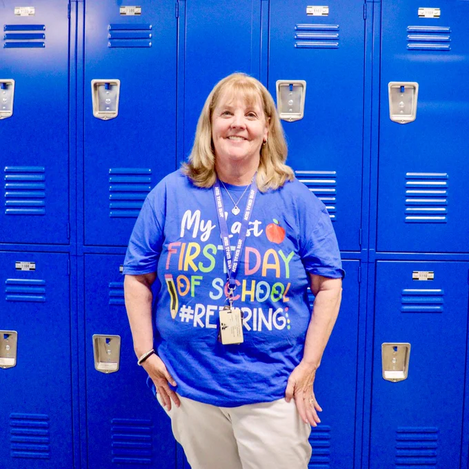 An AMS teacher smiles while wearing a t-shirt that reads "My last first day of school #Retiring"