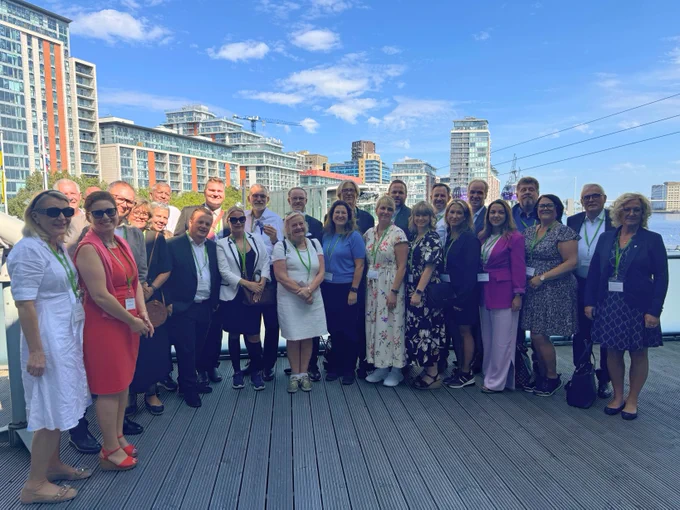 A photo taken outside City Hall of representatives from Salar standing together. Andrew Boff AM and Unmesh Desai are standing at the back, halfway between the right-hand edge and the centre from the group's perspective. The sky is bright, with only a few thin clouds above the group. There are multi-storey buildings in the background, and cable car cables are visible behind the group, to the left from their point of view.