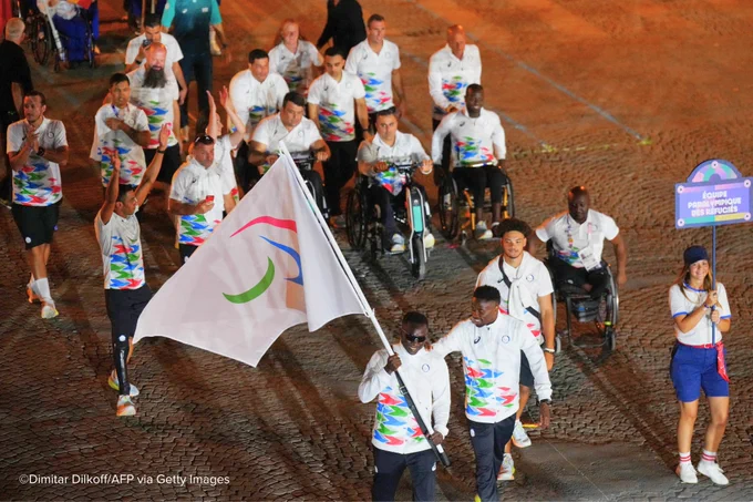 A group of Paralympic athletes, some in wheelchairs and others walking, participate in an event, marching on a cobblestone path. Two athletes at the front proudly carry a white flag featuring the Paralympic symbol in green, red, and blue. A female volunteer on the right holds a sign that reads "Équipe Paralympique des Réfugiés" (Paralympic Refugee Team). The athletes wear white uniforms adorned with vibrant, multicoloured patterns. The scene is lit by warm, ambient lighting, highlighting the camaraderie and spirit of the team.