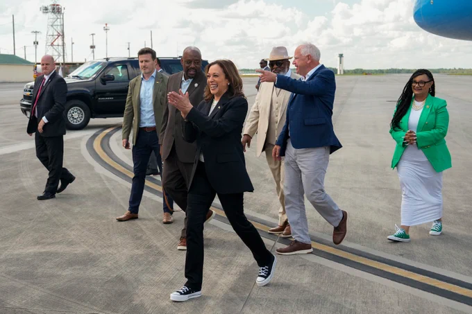 Vice President Kamala Harris and Governor Tim Walz walking on a tarmac with a group of individuals, smiling and gesturing, with an airplane and vehicles in the background.