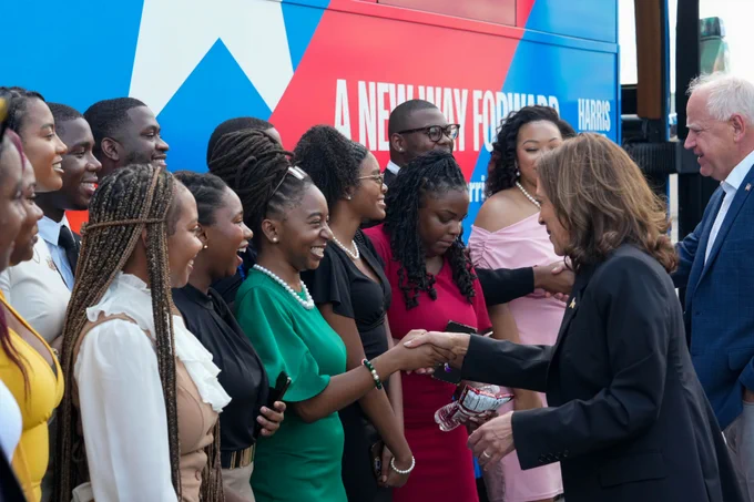 Vice President Kamala Harris and Governor Tim Walz shaking hands with a group of smiling young adults at a public event, with a decorated bus behind them that reads "A NEW WAY FORWARD". 
