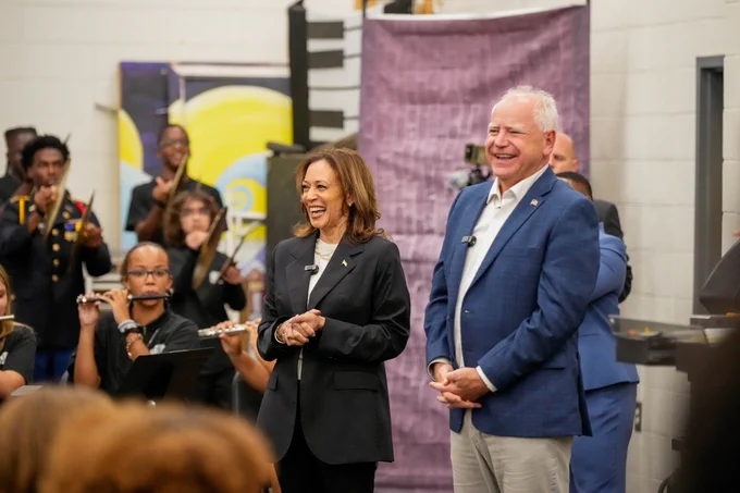 Vice President Kamala Harris and Governor Tim Walz standing together in a room with a school band playing in the background. Both are smiling at the students. 