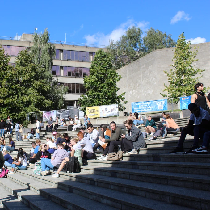 Students sitting on outdoor steps at a university campus on a sunny day, engaging in conversations and enjoying the weather.