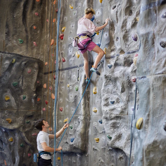 A person climbs an indoor rock wall while another person stands below, handling the safety rope.