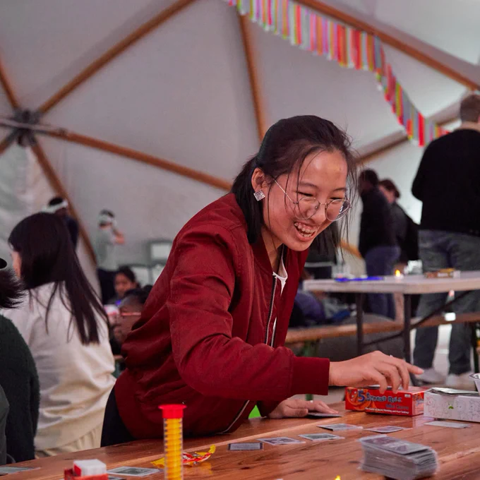 Person in a red jacket smiling while playing a card game at a table in a busy tent with festive decorations.