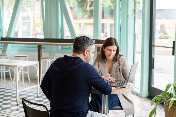 Two people sat at a table, looking at a laptop together.