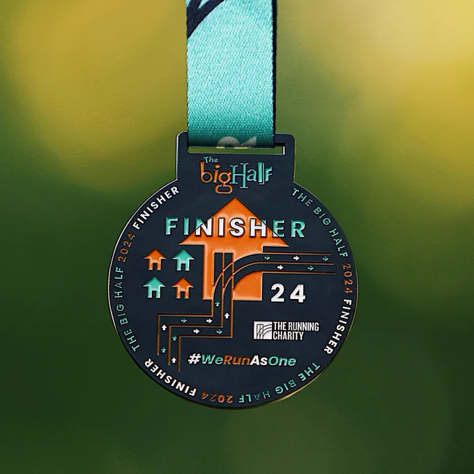A close-up image of the 2024 The Big Half finisher medal, featuring bold orange and blue designs with directional arrows and the text "Finisher" prominently displayed.

The medal is attached to a blue ribbon and set against a green background.