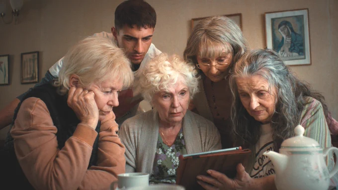 Four elderly women and a young man look intently at a tablet. 