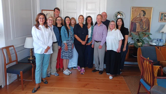 ENNHRI's Board and senior Secretariat staff in a group of 12 look into the camera; they are in a room with wood panel flooring, light grey walls, and a portrait picture of a woman in an expressionist style hangs on the wall on the right-hand side. There is also a plant and numerous pieces of furniture. 