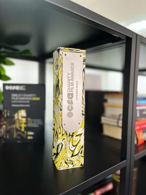 A rectangular block award sits on a black shelving unit with blurred books and a plant in the background. The award is swirled black and yellow with a silver plaque on the front that reads ‘CFA Charity Film Awards. Winner 2025’.
