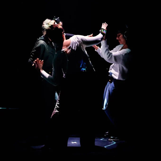 A group of 3 people in a dark environment forming a circle and raising their arms with their hands touching at the fingertips. Two of them (audience members) are wearing headphones and are smiling. The third one (a female dancer) is wearing a phone on their arm displaying a colourful interface. Image credit: Romain Tissot