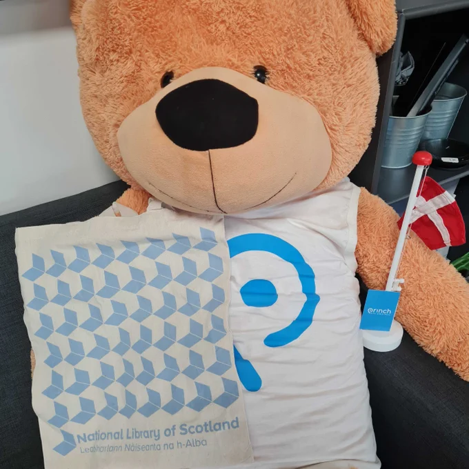 A teddy bear wearing a 'Princh' branded t-shirt sits on a chair with a baby blue National Library of Scotland totebag on one shoulder, and a Danish flag by the other.