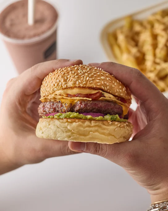 Person holding a nacho cheeseburger with sesame bun, visible in the foreground. In the background, a tray of fries and a chocolate milkshake.