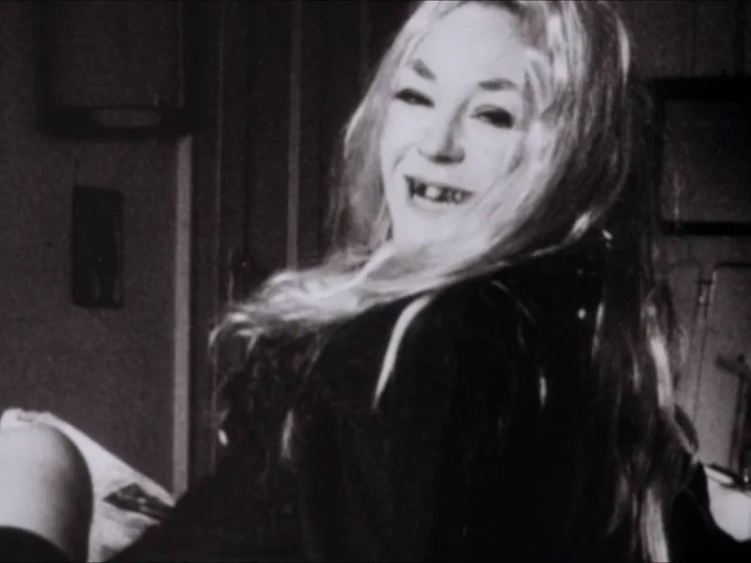 A woman with decayed teeth and long blonde hair (Elsie Downey) turns towards the camera, grins, and stares at the lens in a freeze-frame.