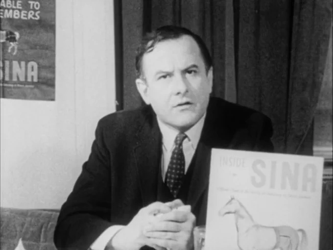 A middle-aged man (Alan Abel) in a black suit and tie stares into a camera with a serious expression. He sits at a desk with a pamphlet in front of him with a cover that reads "Inside SINA" and displays an illustration of a horse wearing pants.