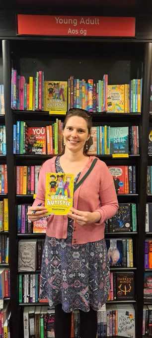 Me standing in front of the kid's bookshelves in Waterstones holding my book "Being Autistic (and what that actually means)"