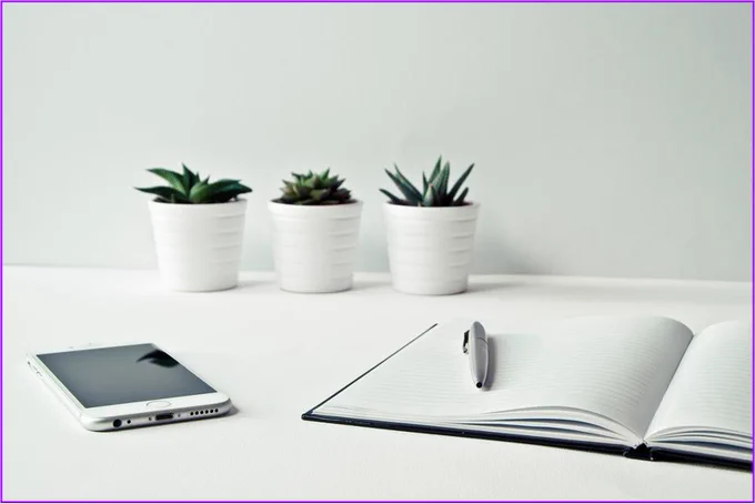 The main colour of image is white.

At the rear of the image is three smooth white planters, each with a green succulent plant in it. The planters are placed on a white surface against a white wall.

In the foreground of the image on the left hand side is a white mobile phone with a black screen.

The foreground of the right hand side of the image contains an open A4 size notebook - there is no writing on the pages. 
A pen is placed on top of the notebook pages on the left of the open book.