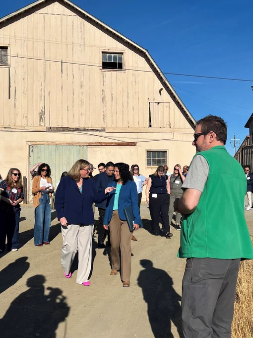 Group of people listening to a speaker wearing a green vest outside a large barn.