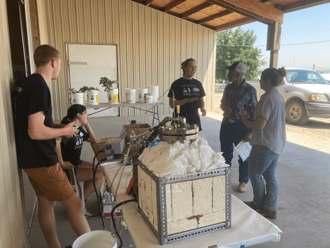 Five people stand around a white and metal kiln that sits on a table on a porch.