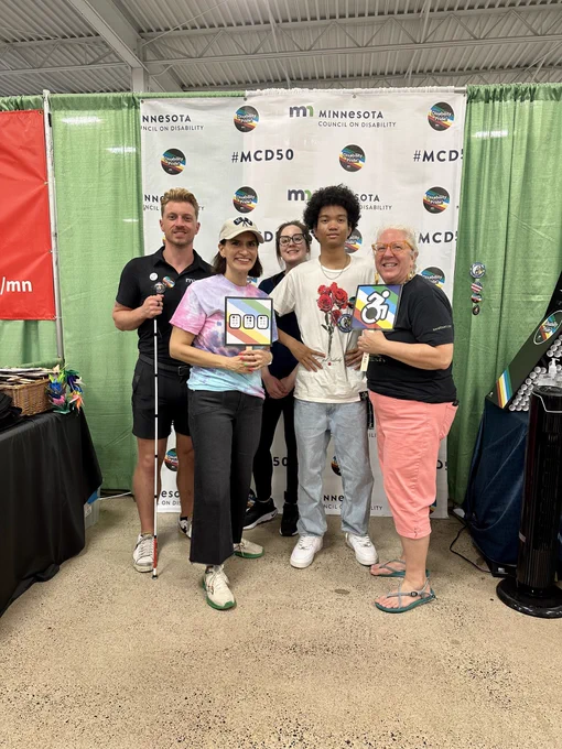A group of five people are standing inside the MCD booth at the State Fair. They are smiling and two people are holding disability related signs. 