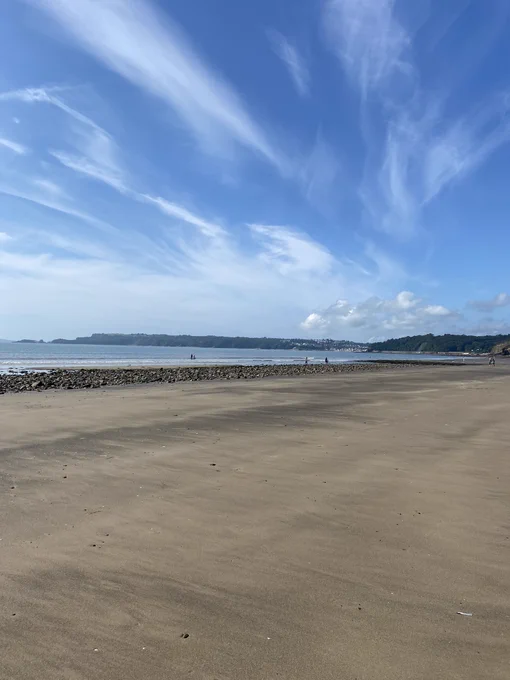 Blue skies and white clouds ahead of Amroth beach. The beach has flat, golden sands and there are green trees in the background.
