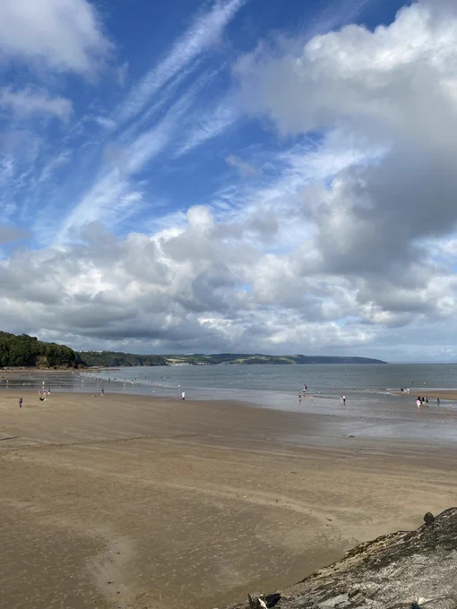 Blue skies and white clouds over Saundersfoot beach. The sand is flat and golden and the water is blue and calm. There are people dotted around the beach and there is greenery in the background.