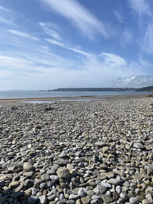 A pebbled Amroth beach with gold sands ahead and the blue sea before it. The skies overhead are blue with white clouds. You can see land in the background.