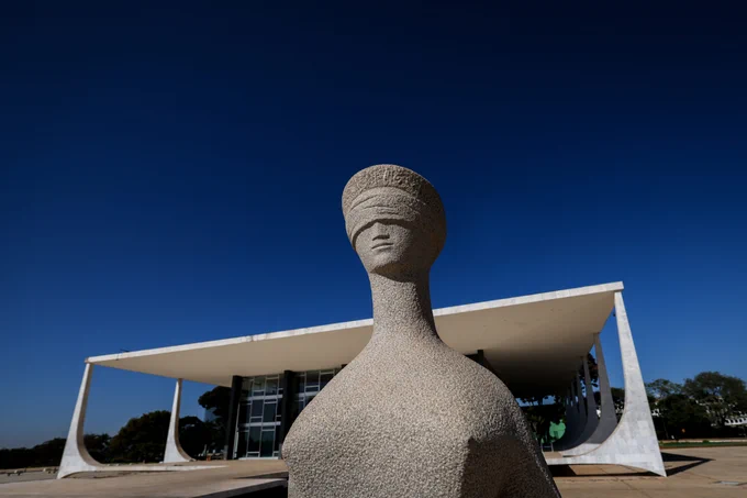 Fotografia horizontal da estátua da Justiça, situada na Praça dos Três Poderes, em primeiro plano. Em segundo plano, o edifício-sede do Supremo Tribunal Federal, e ao fundo, vegetações e folhagens.