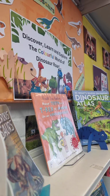 Book display of children's dinosaur books with paper dinosaur crafts on the shelf and above the books.