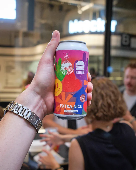 A person holds up a colourful can of "Extra Nice" beer in a crowded restaurant.