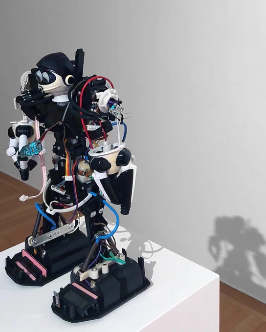 The photo shows a 40 cm high humanoid robot standing on a white pedestal in a gallery. The robot has a mechanical and exposed design, with various visible components such as gears, wires, and circuit boards. Its limbs and torso are made up of a combination of black, white, and metallic parts, giving it a somewhat skeletal and industrial appearance. The robot's head is equipped with two circular eyes and a helmet. The background is plain, with a shadow of the robot cast on the wall behind it, adding depth to the image.