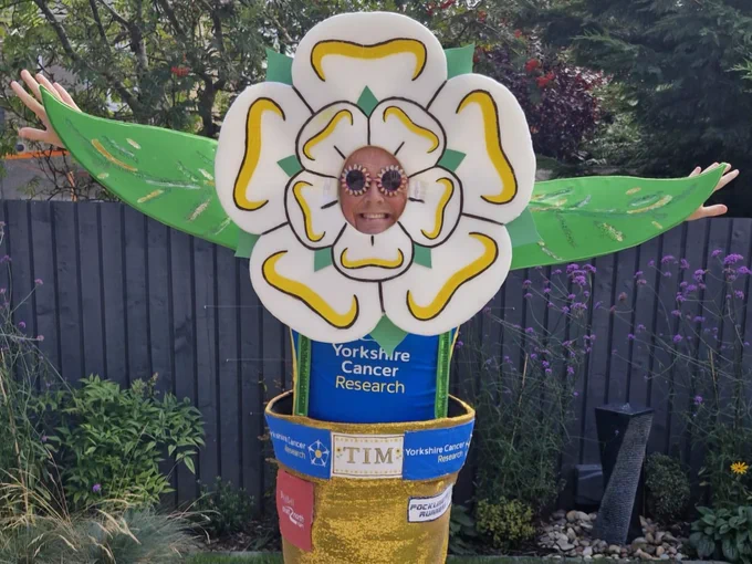 Tim is wearing a large, decorative Yorkshire Rose. His face is visible in the middle of the flower, and he is wearing sunglasses and smiling. Tim's arms are outstretched, showing the two leaves he has created on either side. He is wearing a Yorkshire Cancer Research running vest.