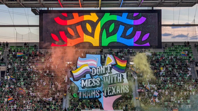 Photo shows a TIFO displayed infront of the supporters section in Q2 stadium. The artwork is of an outline of the state of Texas filled with the trans flag colors overlayed the state flag. The text is written in three lines on top of the state and reads “DON’T MESS WITH TRANS TEXANS”. The text is is white with a black outline, except the word “TRANS” is filled in with the trans flag. On the left side of the word “DON’T” is a trans flag and a nonbinary flag. On the right side of the word “DON’T” is an intersex inclusive pride flag.
