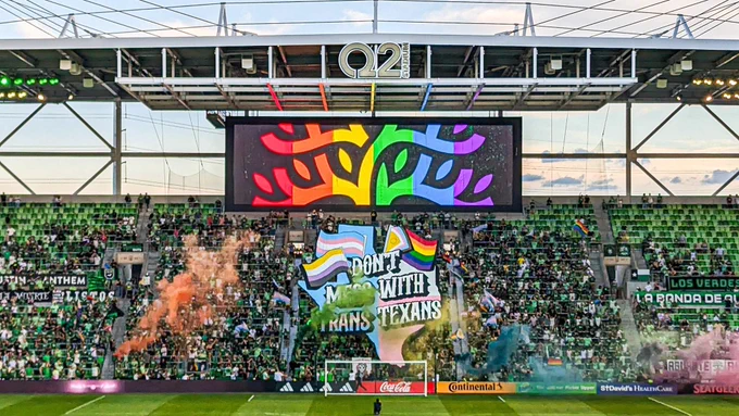 Photo shows a TIFO displayed infront of the supporters section in Q2 stadium. The artwork is of an outline of the state of Texas filled with the trans flag colors overlayed the state flag. The text is written in three lines on top of the state and reads “DON’T MESS WITH TRANS TEXANS”. The text is is white with a black outline, except the word “TRANS” is filled in with the trans flag. On the left side of the word “DON’T” is a trans flag and a nonbinary flag. On the right side of the word “DON’T” is an intersex inclusive pride flag.