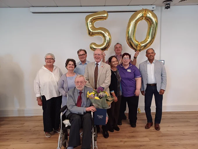 Mike surrounded by friends, family, and staff from the hospital he received his transplant from. They are in a hall with gold balloons in the shape of '50' blown up behind them. 