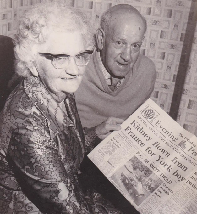 Two elderly individuals seated, holding up a newspaper titled 'Evening Press'. The newspaper's visible headline reads, "Kidney flown from France for York boy".