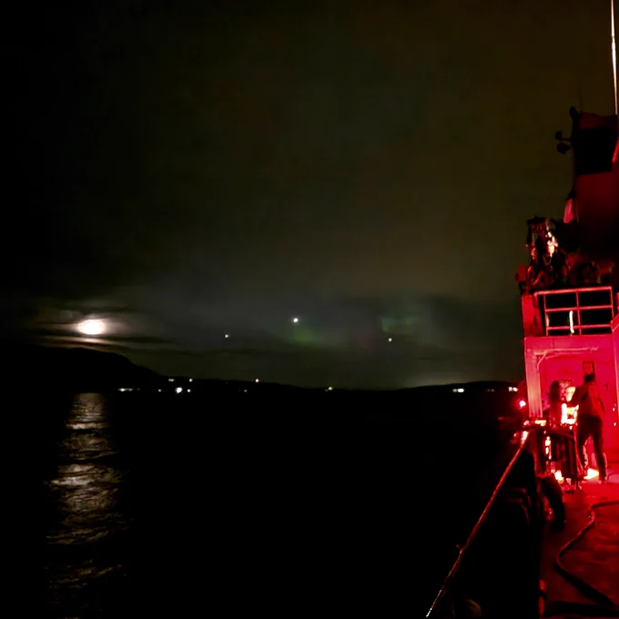 Photograph captured at sea at night with the side of a boat under red illumination on the right side of the image. On the left side is the moon and it's reflection in the water and in the middle green northern lights play on the sky.